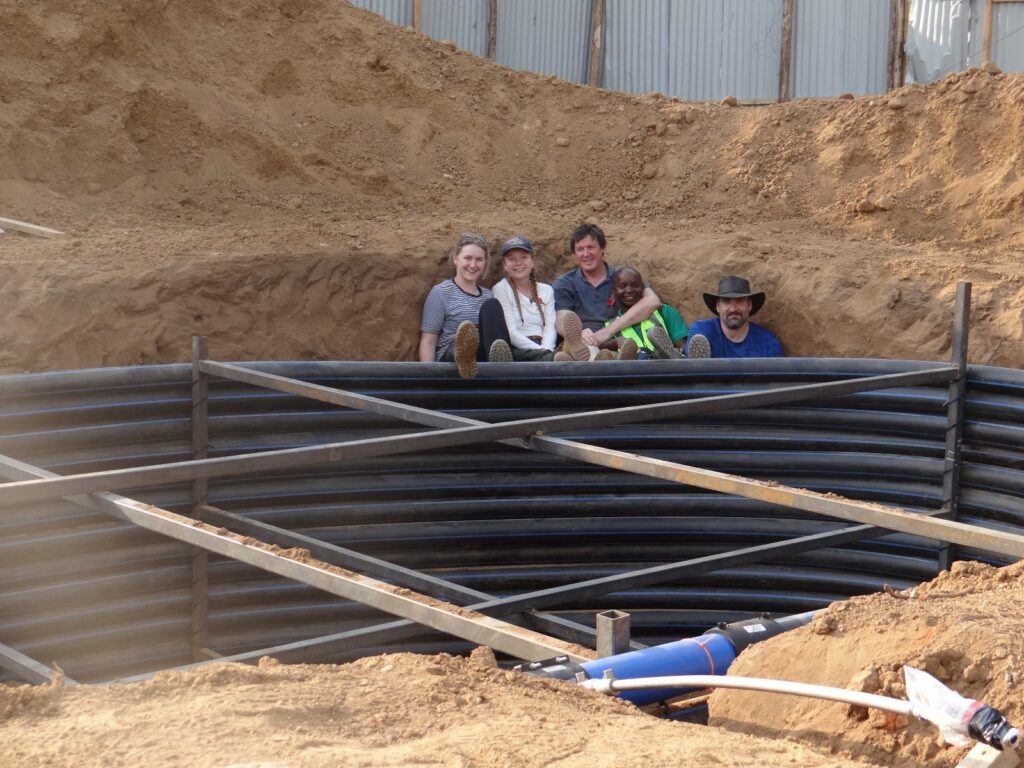 Female and male engineers and a male renewable energy engineer from Malawi sat inside a pit where a hydrogen storage tank is being installed. Large pile of earth in the background, large coiled pipes and bracing structure in the foreground